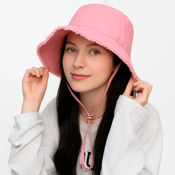 Une femme porte un joli bob chapeau rose avec ficelle et elle tient les bords avec ses mains pour un côté plus fun sur un fond blanc