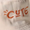 Zoom sur une inscription brodée en orange "so cute" d'un bob chapeau