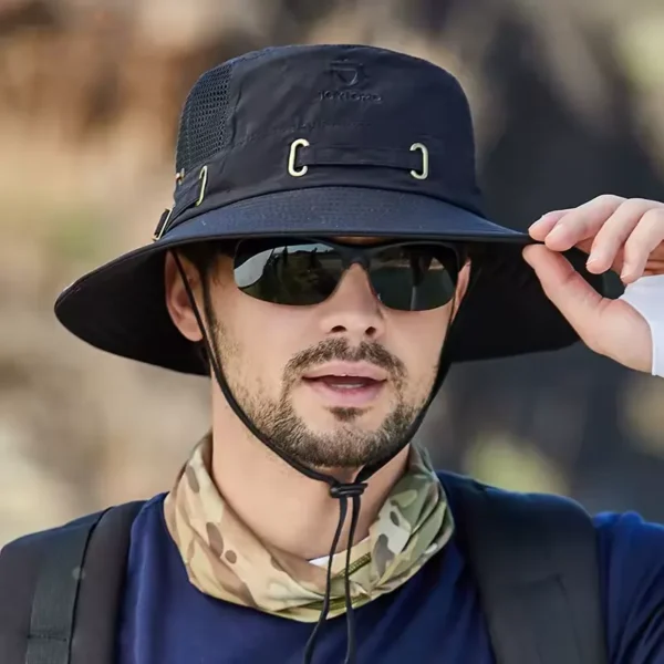 Un homme de face avec des lunettes noires de soleil porte un joli chapeau bob pêche couleur noire sur la tête sur un décor de nature flouté