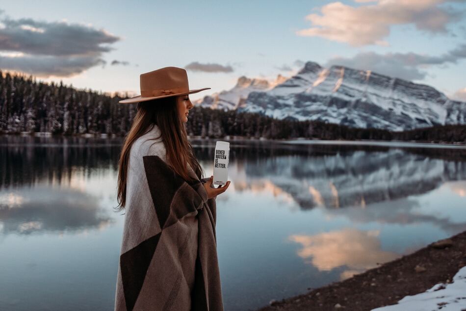Femme avec un chapeau tendance se réchauffe dans son plaid au bord d'un lac en hiver