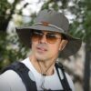 Un homme style explorateur porte un chapeau aventurier avec des grosses lunettes dans un décor de nature