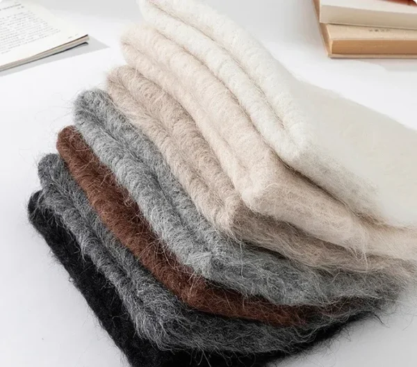 Pile de bonnets en alpaga pliés, montrant différentes teintes naturelles comme beige, gris, marron et noir, soulignant la douceur de la laine d’alpaga.