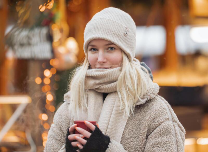 Une jeune femme blonde très élégante et couverte d'un manteau épais et d'une longue écharpe porte un bonnet chaud blanc en laine sur un décor de fêtes de Noël flouté